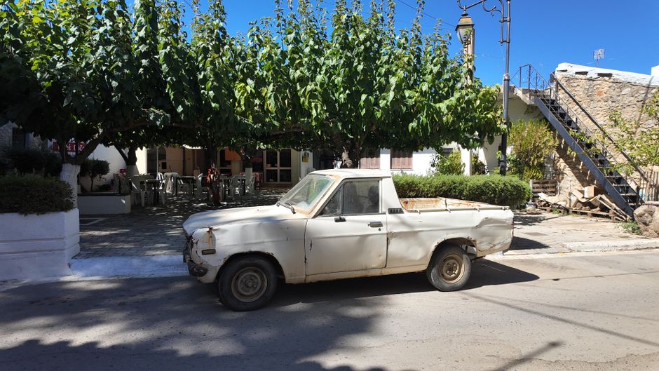 Rusty pickup truck parked on a sunny street in Mochos, Greece, with surrounding trees and buildings.